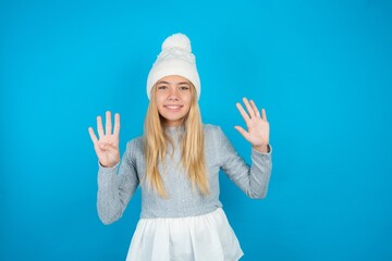 Wall Mural - Teen caucasian girl wearing blue knitted sweater and woolly hat showing and pointing up with fingers number nine while smiling confident and happy.