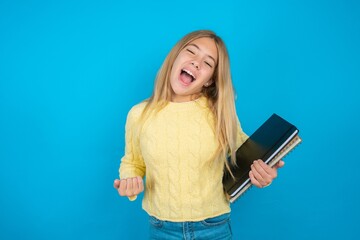 Wall Mural - Beautiful kid girl wearing yellow sweater holding notebook celebrating surprised and amazed for success with arms raised and eyes closed. Winner concept.