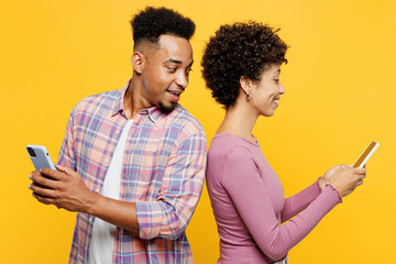 Poster - Side view young cool fun couple two friends family man woman wear purple casual clothes together hold in hand use mobile cell phone stand back to back peep isolated on plain yellow orange background.