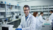 © Studio Nova - Smiling scientist with glasses and a lab coat stands confidently in a laboratory, with shelves stocked with scientific supplies in the background and colleagues working behind him.