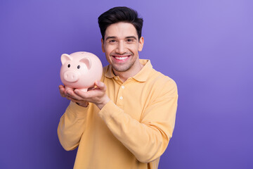 Canvas Print - Photo of cheerful handsome guy wear yellow polo holding piggy money box isolated violet color background