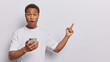 Waist up shot of shocked young African man holds smartphone and points index finger at upper right corner demonstrates something stunning dressed in casual t shirt isolated over white background