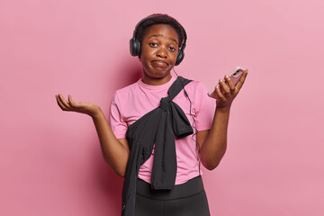 Hesitant dark skinned woman dressed in casual t shirt leggings sweatshirt tied over body holds mobile phone in hand shrugs shoulders with doubt goes in for sport regularly isolated on pink background