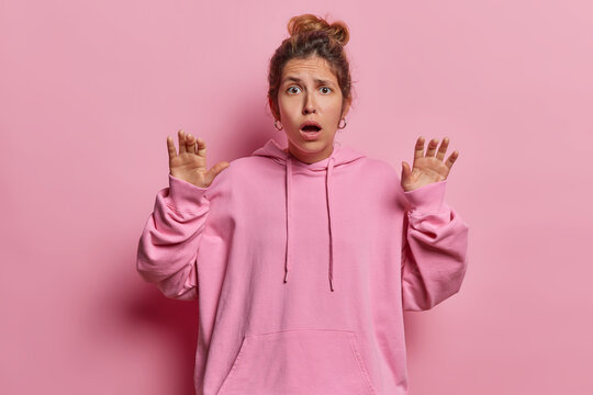 Scared young woman gasps from fear keeps hands raised up holds breath from amazement dressed in casual hoodie poses against pink background reacts to something teerrible. Human reactions concept