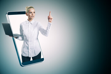 Aufkleber - Attractive young european businesswoman inside smartphone holding laptop and pointing up on gray background with mock up place. Internet communication, online video messaging and remote work concept.