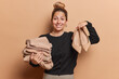 Waist up shot of pleased young European woman holds neatly folded stack of rich brown clothes and high heeled shoes suggests to buy it on sale wears black jumper isolated over brown background