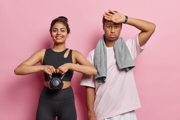 Wall Mural - Sporty lifestyle. Studio shot of young fit happy smiling European girl using weight and tired African american guy wearing tracksuits after doing physical exercises to be healthy and in good shape