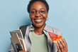 Horizontal shot of cheerful African girl with short dark hair smiles plesantly holds piece of pizza textbooks and cellphone enjoys studying day dressed casually isolated over blue background