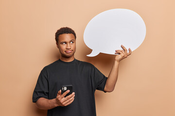 Wall Mural - People and emotions. Studio waist up of young darkskinned man in black t shirt holding smartphone and white speech bubble with blank space for your advertisement standing in centre on beige background