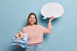Happy young European woman with long hair smiles broadly wears casual jumper and jeans holds folded clothes and sandals holds blank speech bubble for your promotion isolated over blue background