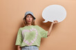 Photo of surprised European woman wears panama and casual t shirt holds white blank speech bubble for your advertising content poses against brown background. People idea and promotion concept