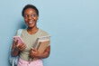 Student lifestyle cocnept. Indoor photo of young happy smiling African american lady standing on left isolated on blue background wearing olive t shirt and pink jeans with blank space for promotion