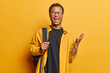 Photo of dark skinned man keeps hand raised up and exclaims loudly carries rucksack on shoulder wears spectacles and casual shirt isolated over yellow background. People and emotions concept