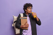 Worried curly haired Hindu student drinks fresh orange juice and carries spiral notepads wears spectacles and black shirt carries rucksack on shoulder poses over purple background. Studying lifestyle