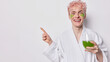 Horizontal shot of pink haired man dressed in domestic robe holds glass of green fresh juice or smoothie undergoes beauty procedures points index finger on empty space isolated over white background