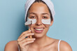 Positive young European woman applies beauty patches and uses face roller for skin care smiles happily enjoys beauty procedures wears towel after taking shower isolated over blue background.