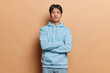 Horizontal shot of handsome confident Asian man with dark hair keeps arms folded feels self confident wers blue sweatshirt and jeans isolated over brown background listens something attentively