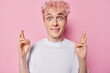 Horizontal shot of pink haired freckled man keeps fingers crossed for having luck bites lips looks surprisingly at camera wears casual white t shirt poses indoor makes cherished wish. Body language