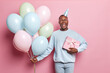 Horizontal shot of emotional dark skinned man holds bunch of inflated balloons and wrapped present exclaims loudly celebrates birthday isolated over pink backgroun. People and holiday concept