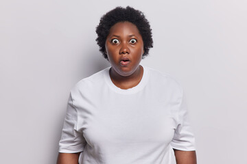 Wall Mural - Indoor shot of stunned African woman with curly hair stares bugged eyes cannot believe in shocking news feels amazed dressed in casual jumper isolated over white background. Human reactions concept