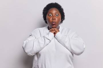 Wall Mural - Waist up shot of dark skinned plump adult woman amazed by smth standing speechless keeps hands under chin lips rounded dressed in casual sweatshirt poses against white background gasps startled