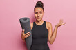 Hesitant doubtful Latin sportswoman has healthy body habits holding rolled fitness mat shrugs shoulders with doubt wears black tracksuit isolated over pink background. Sporty lifestyle concept