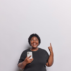 Vertical shot of cheerful dark skinned young woman holds smartphone and points index finger overhead shows free space for advertisement wears casual black t shirt isolated over white background.