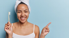 Oral Hygiene Concept. Cheerful Latin Woman Smiles Gladfully Holds Wooden Toothbrush And Points Index Finger Aside On Copy Space Gives Advice How To Care About Teeth Isolated Over Blue Background
