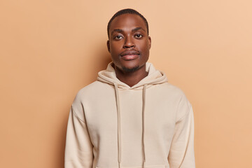 Horizontal shot of handsome dark skinned African man with small beard full lips big eyes looks directly at camera dressed in casual sweatshirt poses against brown background. Human face expressions