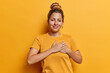 © WHstudio Leushin N - Horizontal shot of pretty young woman presses hands to heart expresses gratitude expressing appreciation and kindness smiles happily dressed in casual t shirt isolated over yellow background