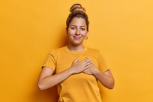 Horizontal Shot Of Pretty Young Woman Presses Hands To Heart Expresses Gratitude Expressing Appreciation And Kindness Smiles Happily Dressed In Casual T Shirt Isolated Over Yellow Background