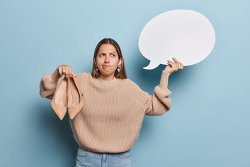 Wall Mural - Horizontal shot of thoughtful long haired European woman holds high heeled shoes and communication bubble thinks about selling them wears knitted jumper and jeans isolated over blue background.