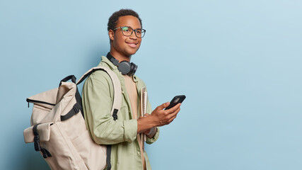 Wall Mural - Horizontal shot of handsome dark skinned male student holds smartphone in hands and carries big rucksack holds textbooks wears spectacles and casual shirt isolated over blue background copy space