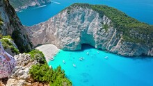 Aerial view of a beautiful woman looking at Navagio Pebble Beach with a shipwreck and turquoise water. Female tourists enjoying summer vacation at the cove reached by boats in Zakynthos, Greece.