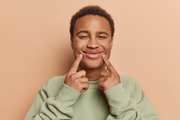 Wall Mural - Positive dark skinned young man keeps fingers near corners of lips forces cheerful smile tries to forget about all problems and be happy dressed casually isolated over brown studio background