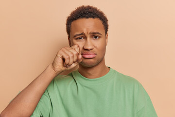 Wall Mural - People negative emotions concept. Studio portrait of upset African american man standing in centre isolated on beige background looking as if crying and wiping tears holding fist under left eye