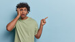 People emotions concept. Studio waist up of young surprised Hindu male standing on left isolated on blue background pointing at blank space for your promotion wearing olive t shirt looking straight