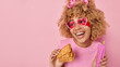 Positive curly haired woman wears trendy heart shaped sunglasses holds waffle toothbrush and toothpaste concentrated aside applies beauty patches under eyes isolated over pink studio background