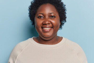 Portrait of cheerful dark skinned body positive African woman smiles gladfully gets good news dressed in casual jumper looks directly at camera isolated over blue background. People and emotions