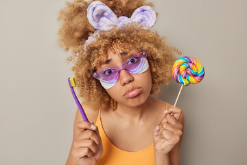Wall Mural - Dental care concept. Indoor shot of young beautiful African american female standing in centre isolated holding lollipop and purple toothbrush in yellow t shirt upset can't eat candy wearing glasses