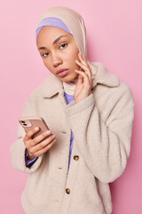 Wall Mural - Photo of Muslim woman has serious expression touches face holds mobile phone in hand wears hijab and coat focused directly at camera isolated over pink background. People and technology concept
