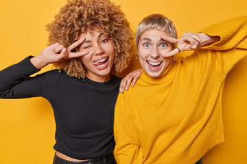 Poster - Horizontal shot of cheerful two women make peace gesture over eyes smiles gladfully winks and sticks out tongue dressed casually isolated over yellow background. Friendship and body language concept