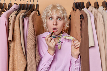 Photo of fair haired European woman stares surprised at camera puts on red lipstick gets ready for business meeting shocked to be late wears elegant clothing surrounded by outfits on hangers