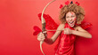 © WHstudio Leushin N - Horizontal shot of emotional female cupid takes aim with her bow and arrow gracefully shooting towards undisclosed targets wears elegant red dress that complements angelic wings isolated on red wall