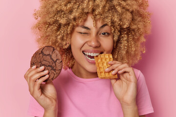 Wall Mural - Positive curly haired young woman eats delicous cookie and waffle winks eye and smiles broadly dressed in casual t shirt has sweet tooth eats favorite desserts isolated over pink background.