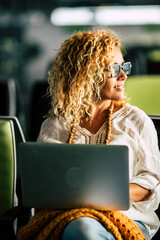 Wall Mural - Adult blonde curly woman work at laptop computer sitting inwaiting room at the airport alone - travel and modern people in smart working remote worker activity with technology and internet