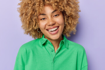 Wall Mural - Portrait of good looking woman with blonde curly hair smiles toothily expresses postive emotions wears earrings and green jumper isolated over purple background. People and happiness concept