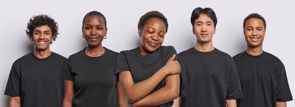 Photo of dark skinned young woman embraces own body keeps eyes closed smiles gently poses among other four people with glad expressions dressed in black t shirts isolated over white background