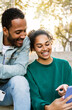 © Xavier Lorenzo - Vertical shot of two young latin american people using smart phone together sitting outdoors. Cheerful african american friends laughing while looking at cellphone screen. Social media concept.