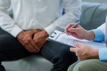 Wall Mural - close-up of male patient people seated, clutching his lower abdomen, while doctor with clipboard sits beside him, possibly discussing prostate health issues.